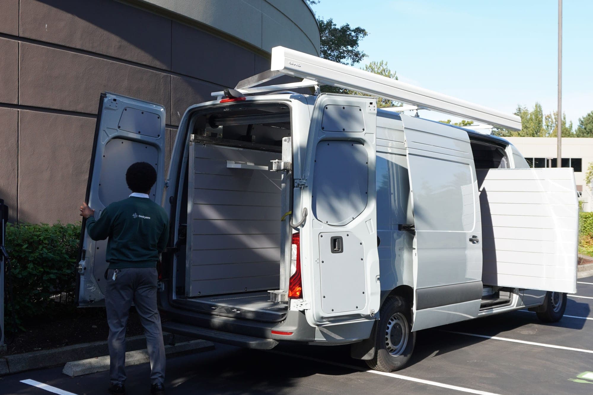 The Mobile Market van in profile with its rear doors open and the awning stowed on the roof, a Denkyem team member opening it up
