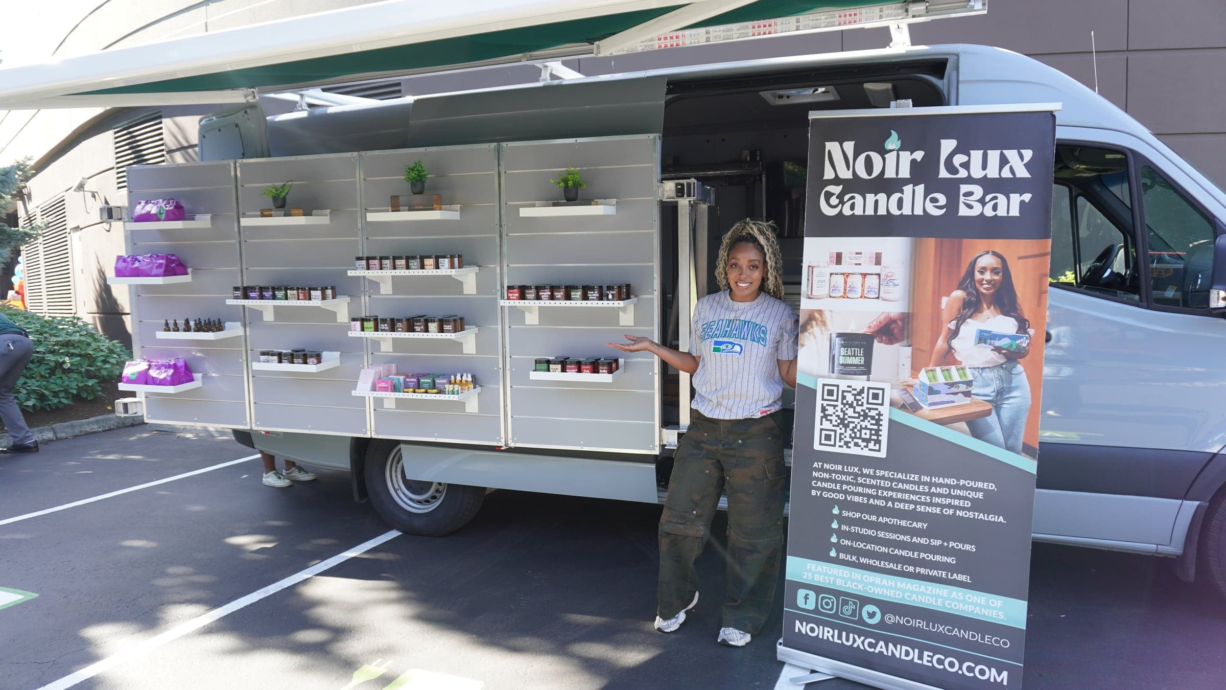 The Mobile Market van open at an event: shelves stocked with candles and skincare, the awning out, and Colina of Noir Lux Candle Bar standing at the open side door
